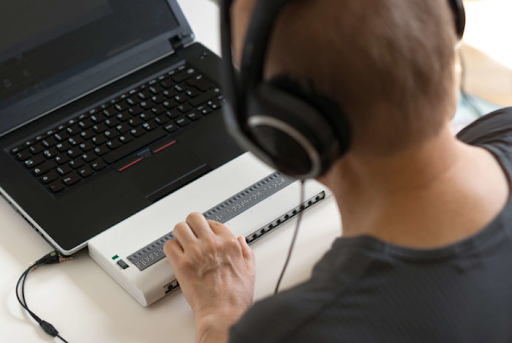 Close up image of a man sitting at a computer with a screen reader attached. He is also wearing headphones.